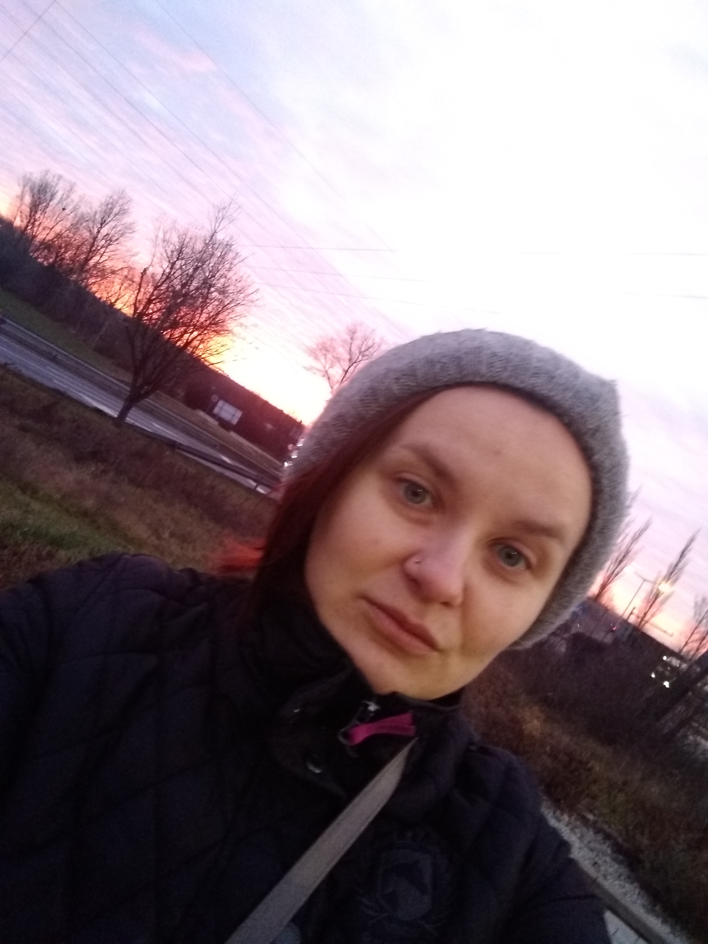 Portret kobiety w szarej czapce na tle zachodzącego słońca i drzew bez liści.