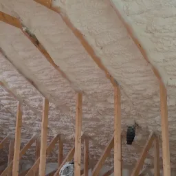 Poddasze z drewnianą konstrukcją krokwiową pokryte grubą warstwą jasnej pianki izolacyjnej, widoczne elementy wentylacji i przewody elektryczne.