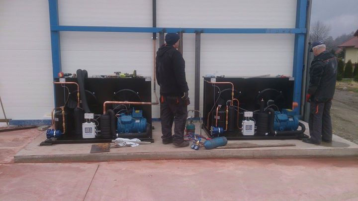 Dwaj mężczyźni instalują dwie identyczne jednostki chłodnicze na betonowej podstawie wewnątrz budynku o białych ścianach z niebieską ramą, widoczne narzędzia i butle gazowe.