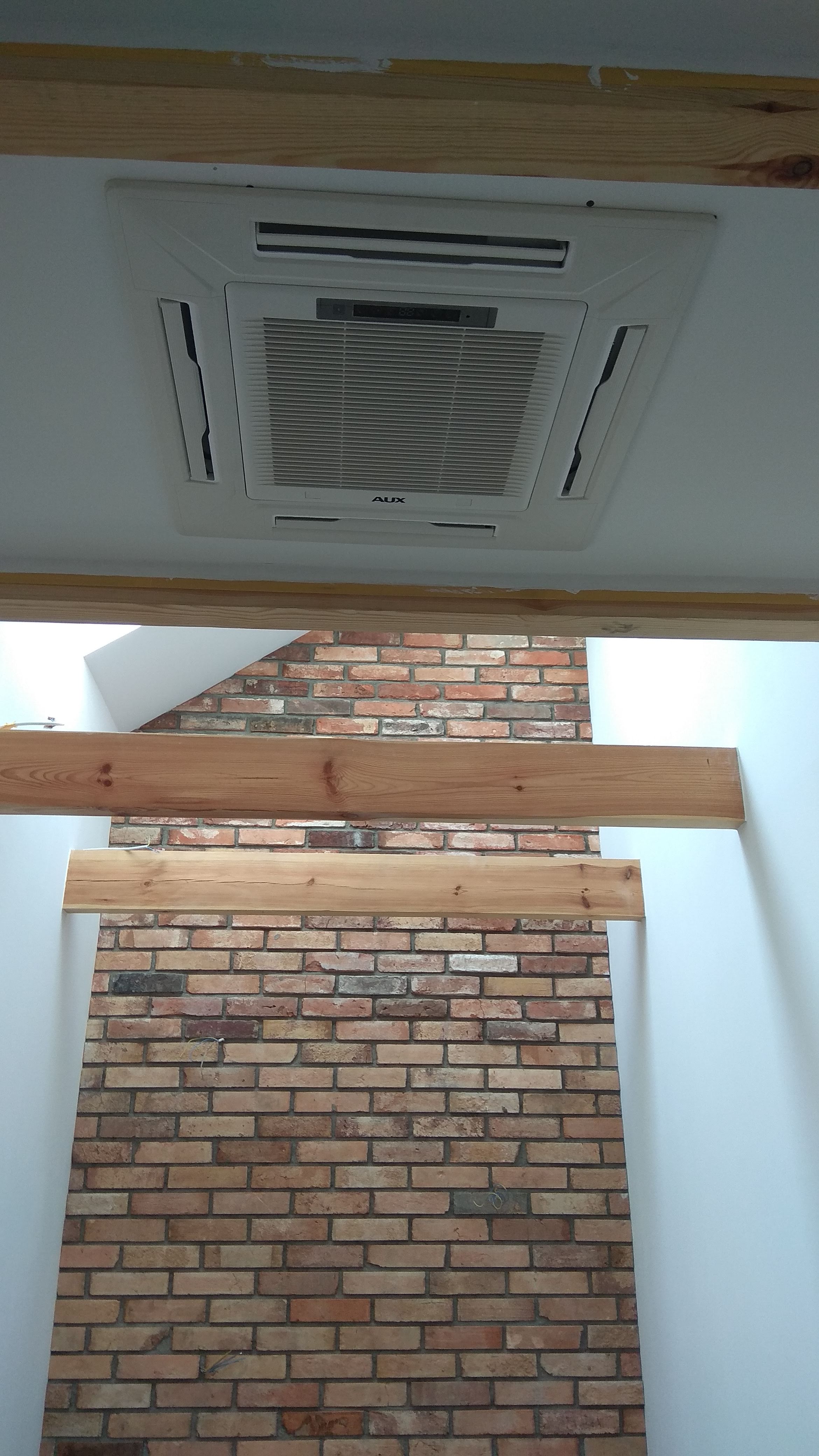 Sufitowa jednostka klimatyzacyjna AUX zamontowana w pomieszczeniu z dekoracyjną ścianą z cegły, widoczna z perspektywy osoby stojącej pod nią.