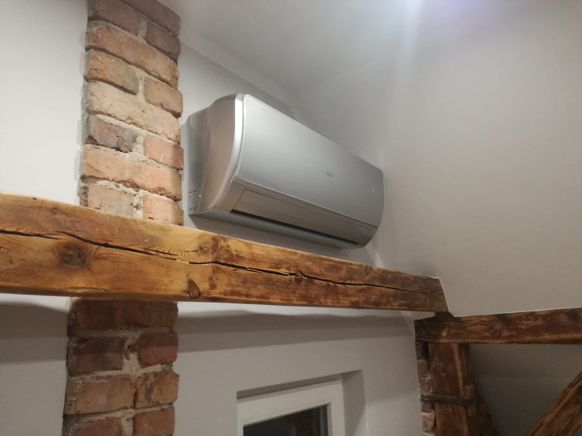 Srebrny klimatyzator ścienny zamontowany w pomieszczeniu z elementami rustykalnymi: ceglana kolumna i drewniana belka stropowa.