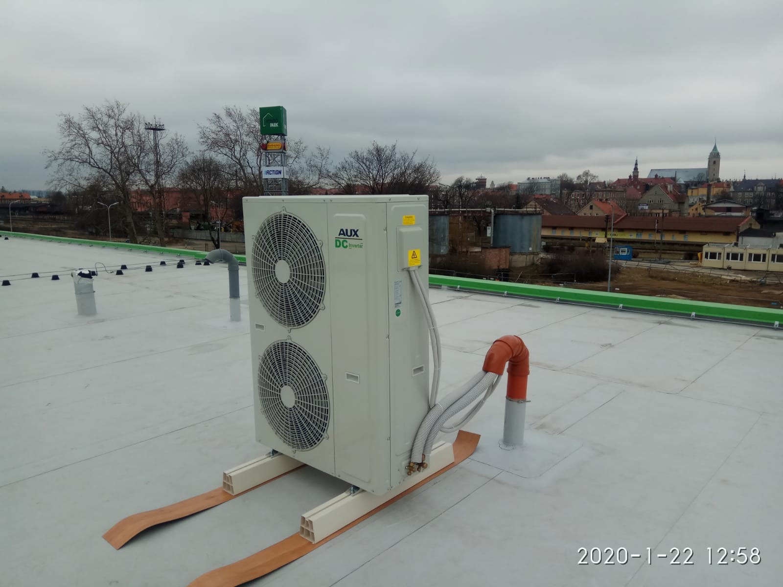 Agregat klimatyzacyjny AUX DC Inverter na płaskim dachu budynku, widoczne przewody, rura wentylacyjna i panorama miasta w tle, pochmurne niebo.