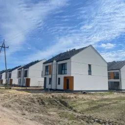 Szereg identycznych, nowoczesnych domów jednorodzinnych z białymi elewacjami i ciemnymi dachami, widoczne na tle błękitnego nieba z chmurami, teren wokół domów jeszcze niezagospodarowany, widoczny...