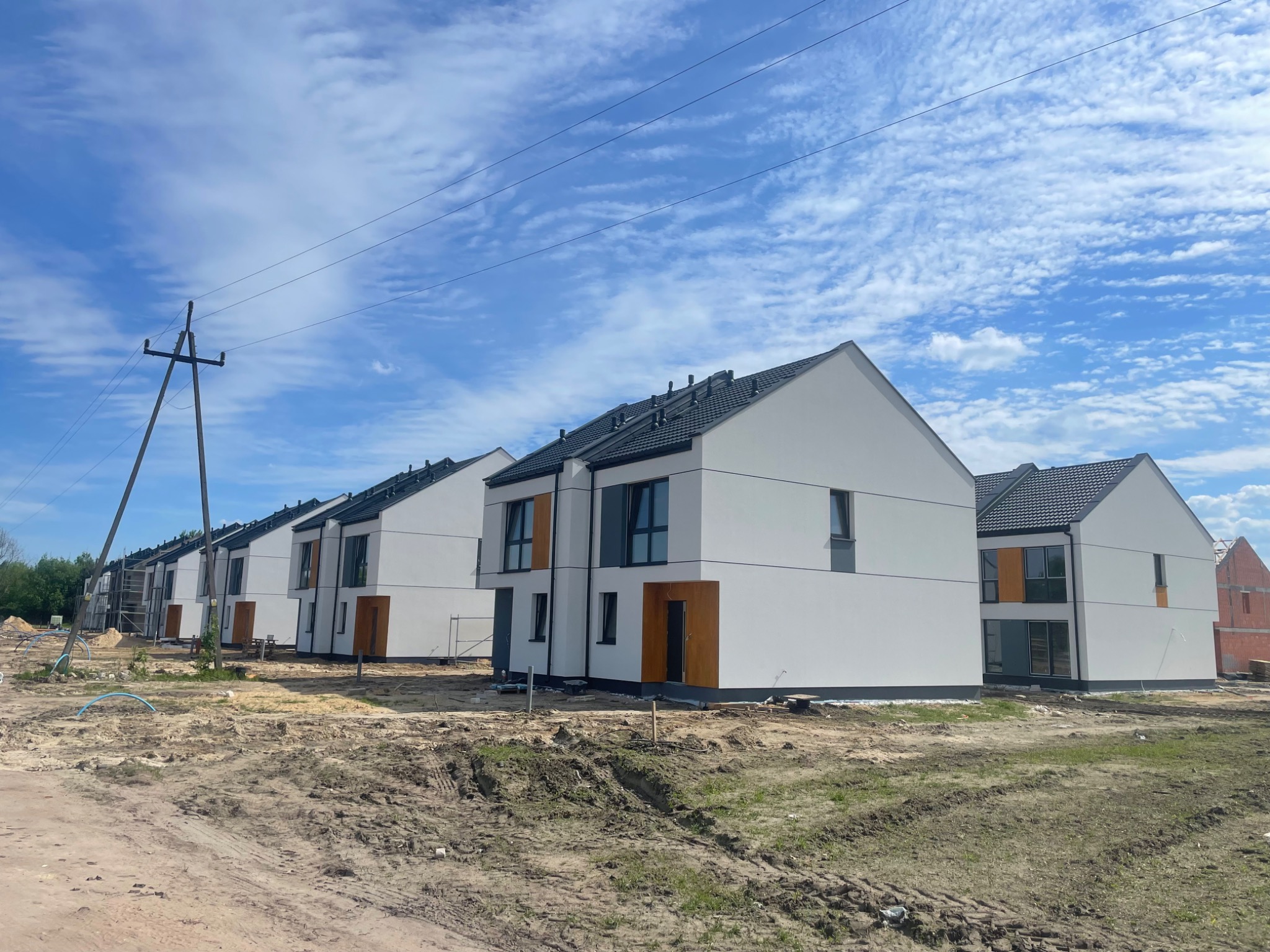 Szereg identycznych, nowoczesnych domów jednorodzinnych z białymi elewacjami i ciemnymi dachami, widoczne na tle błękitnego nieba z chmurami, teren wokół domów jeszcze niezagospodarowany, widoczny...