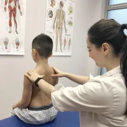 Chłopiec siedzi na niebieskim stole do badań, a kobieta w białym fartuchu kładzie ręce na jego plecach; w tle plakaty z układem mięśniowym i nerwowym człowieka.