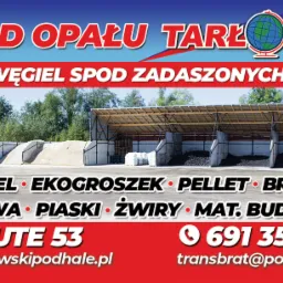 Szeroki kadr na skład opału z zadaszonymi boksami wypełnionymi węglem, pelletem i brykietem, w tle zieleń drzew, na pierwszym planie betonowe przegrody i materiały sypkie, baner reklamowy z nazwą...