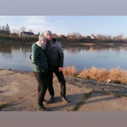 Para w zimowych ubraniach pozuje na tle rzeki i zabudowań w słoneczny dzień. Napis 'Nowy Rok 2020' w górnej części zdjęcia.