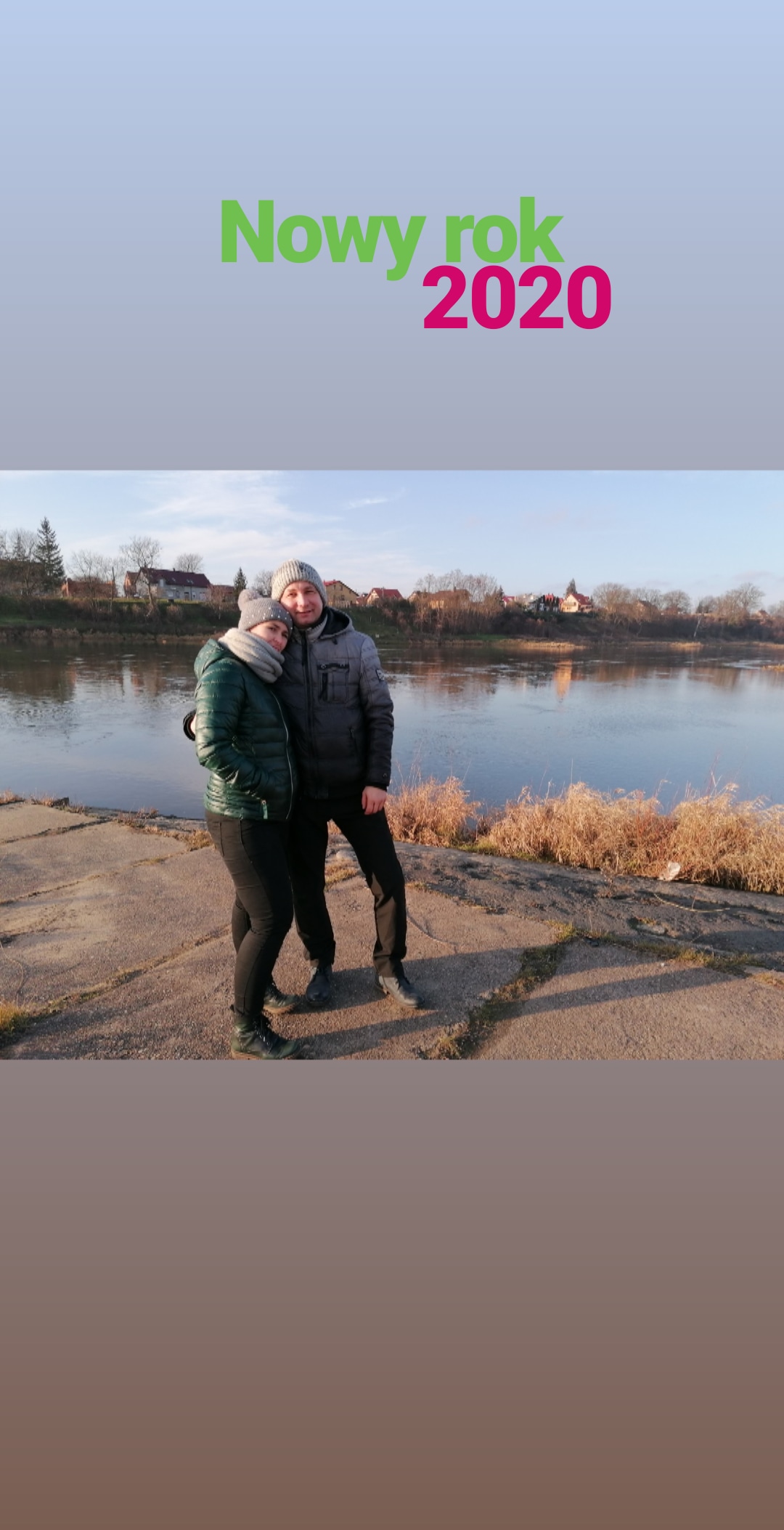 Para w zimowych ubraniach pozuje na tle rzeki i zabudowań w słoneczny dzień. Napis 'Nowy Rok 2020' w górnej części zdjęcia.