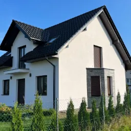 Nowoczesny dom jednorodzinny z białą elewacją, czarnym dachem z dachówki i brązowymi roletami, otoczony zielonym ogrodzeniem z siatki i młodymi tujami.