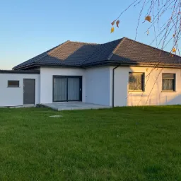 Jednopiętrowy dom z szarą dachówką i białą elewacją w promieniach zachodzącego słońca, widok z zadbanego trawnika.