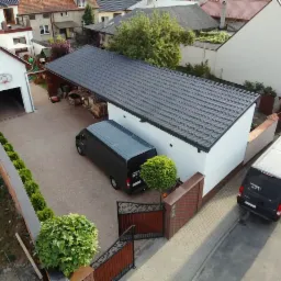 Widok z góry na posesję z dwoma czarnymi samochodami dostawczymi, garażem z koszem do koszykówki i nową, białą elewacją budynku z ciemnym dachem.