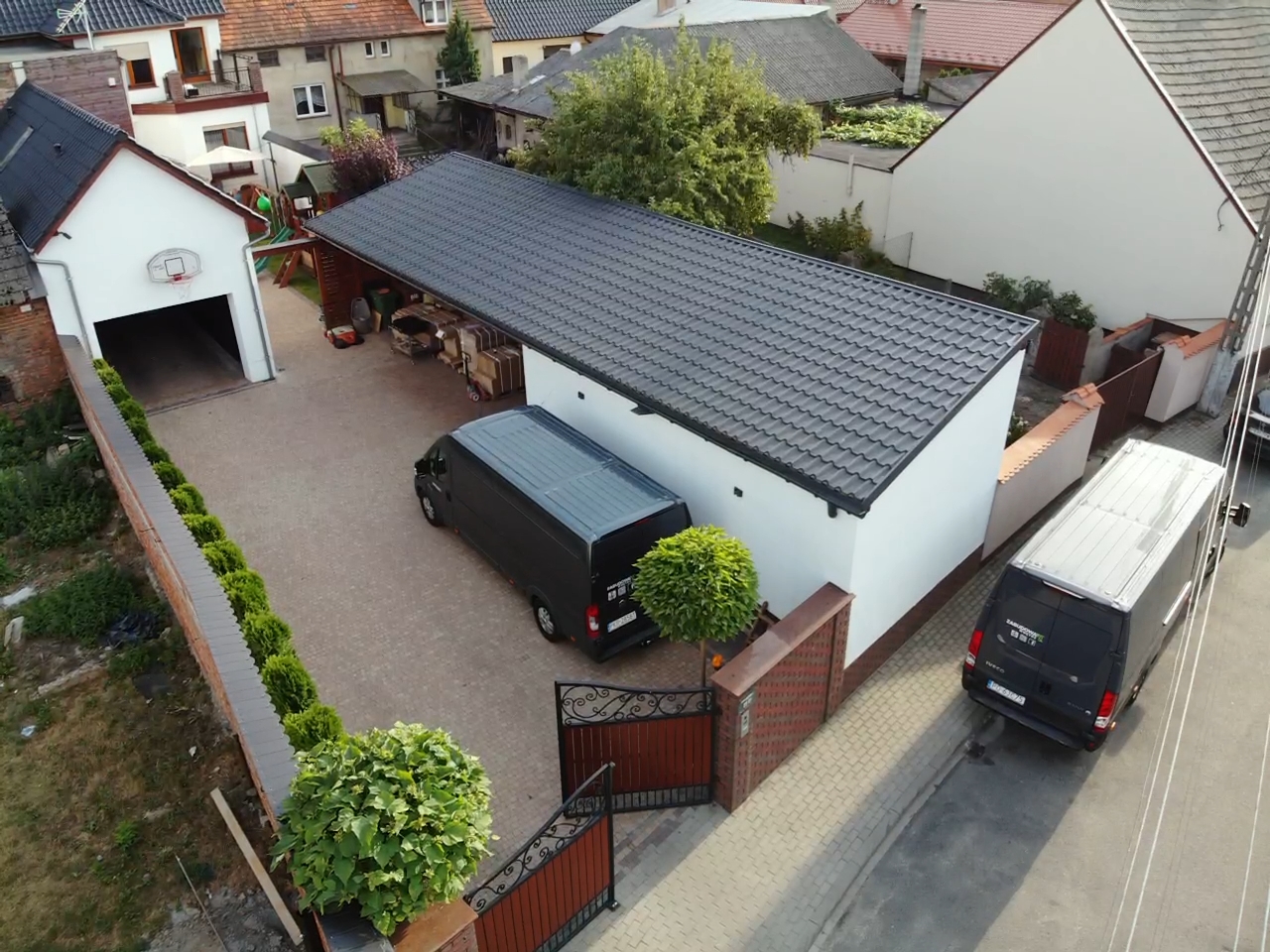 Widok z góry na posesję z dwoma czarnymi samochodami dostawczymi, garażem z koszem do koszykówki i nową, białą elewacją budynku z ciemnym dachem.