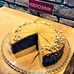 Przekrojony tort orzechowy z karmelem na szklanej paterze, na drewnianym blacie, obok ceglanej ściany z tabliczką 'KIEROWNIK'.