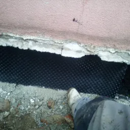 Czarna folia kubełkowa przytwierdzona do fundamentu budynku, widoczny fragment ściany z naprawionymi ubytkami, na dole gruz i stopa w roboczym obuwiu.