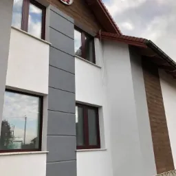 Elewacja domu z elementami dekoracyjnymi: szara wstawka, imitacja drewna, kamienna podmurówka i herb na szczycie ściany.