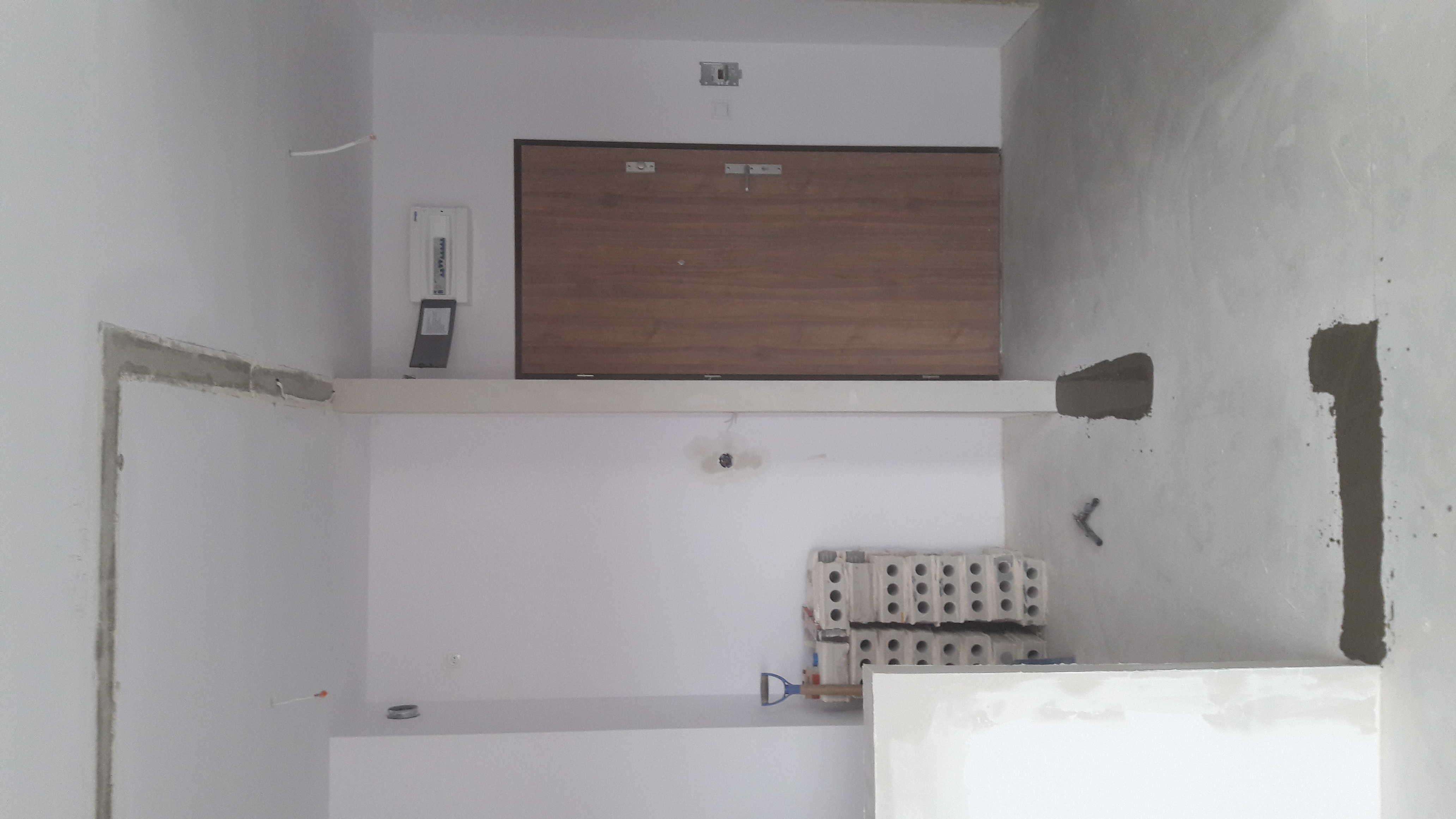 Wnętrze w trakcie prac wykończeniowych: widoczne ściany z surowym tynkiem, przygotowane bloczki, skrzynka elektryczna oraz zamontowane drzwi wewnętrzne z brązową okleiną.