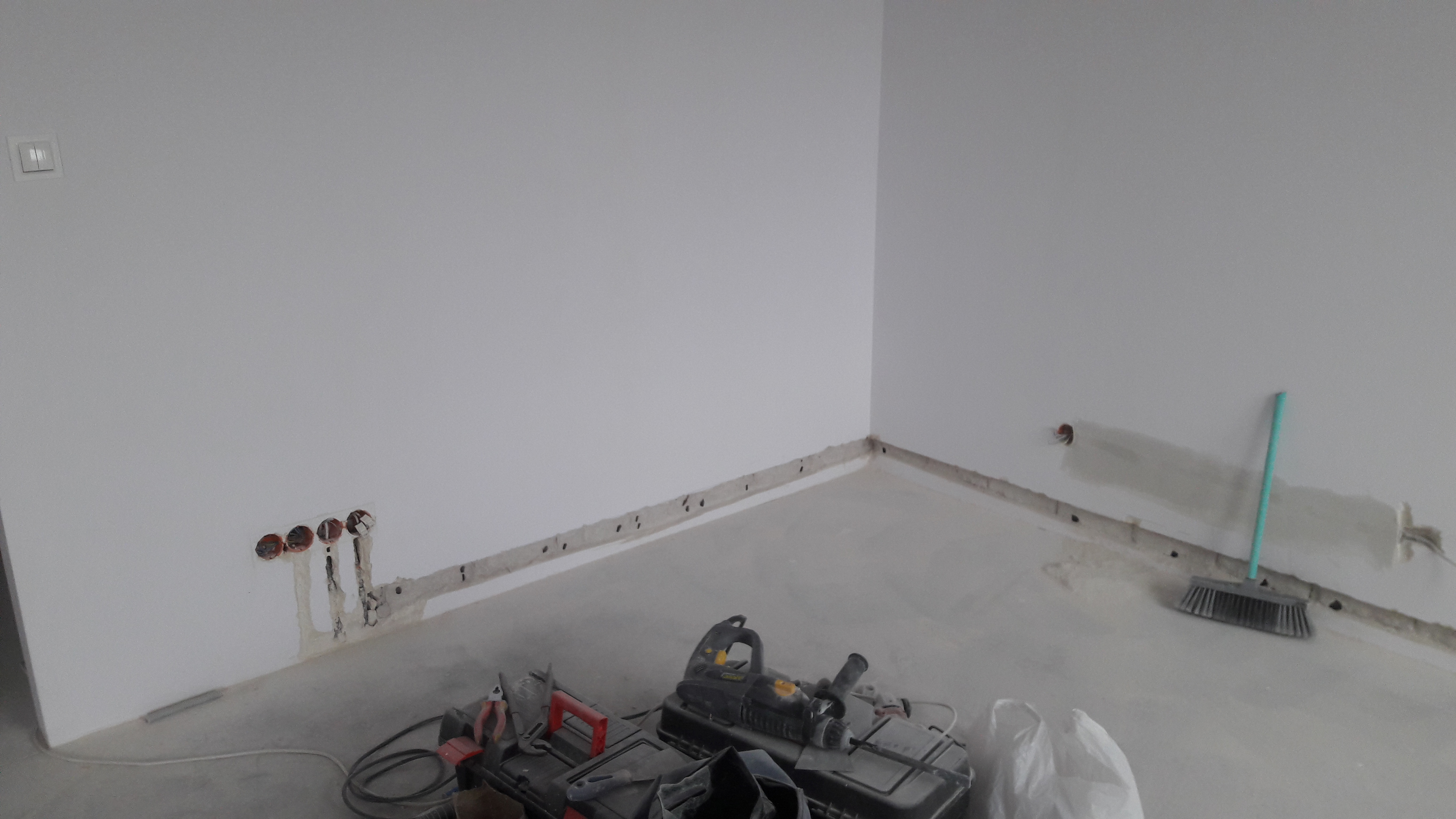 Surowe wnętrze w trakcie prac wykończeniowych: widoczne bruzdy pod instalacje elektryczne w ścianach, narzędzia pracy na podłodze oraz otwory pod gniazdka elektryczne.
