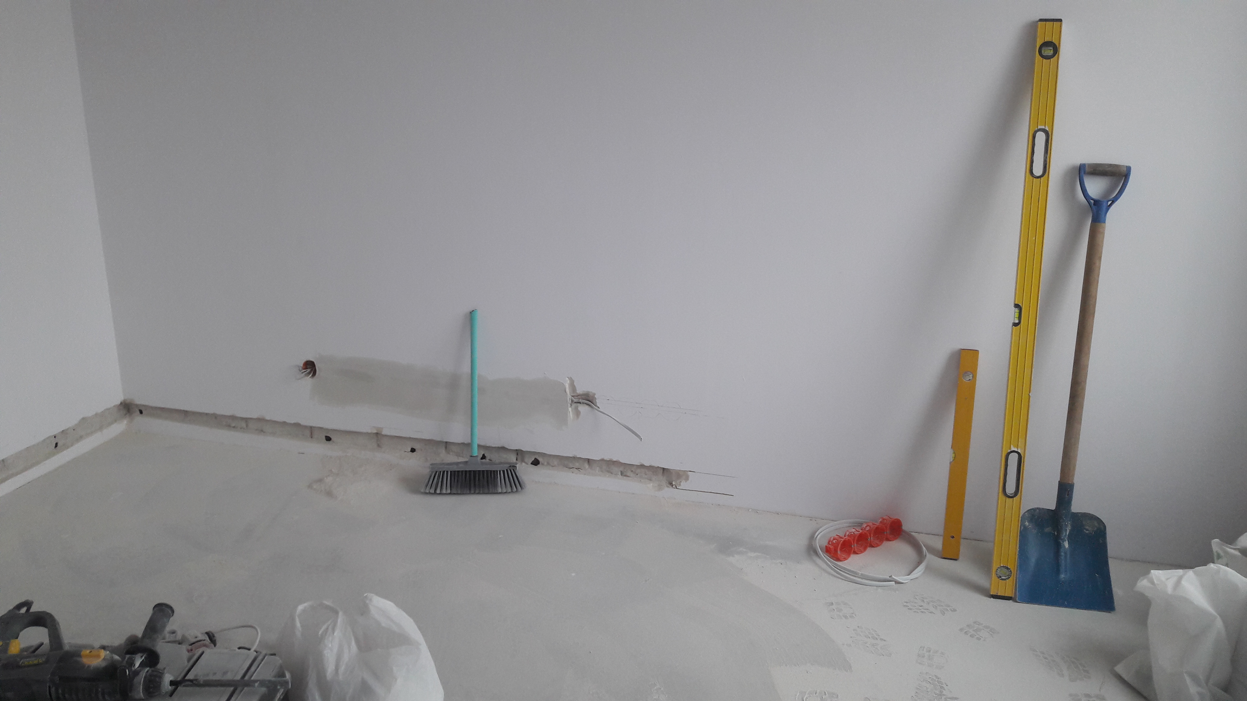 Pokój w trakcie prac, z widocznymi śladami po kuciu w ścianach, szpachlą, przewodami elektrycznymi, narzędziami (poziomice, szpachla) i sprzętem do cięcia płytek na podłodze, z pozostawioną miotłą...