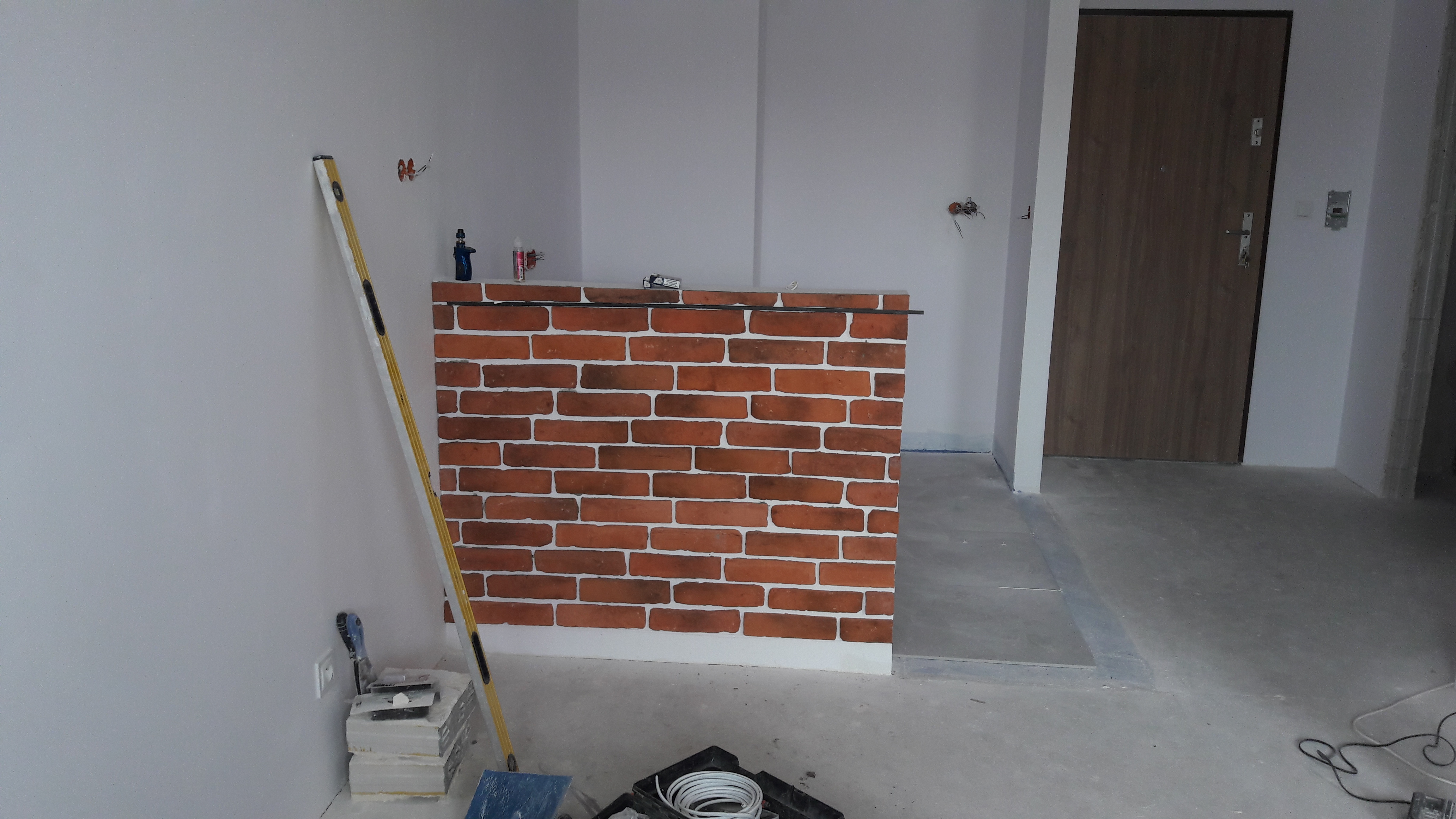 Wnętrze w trakcie prac wykończeniowych: ściana z cegieł klinkierowych, poziomica oparta o ścianę, widoczne narzędzia malarskie i puszki z farbą na tle surowych ścian i podłogi.