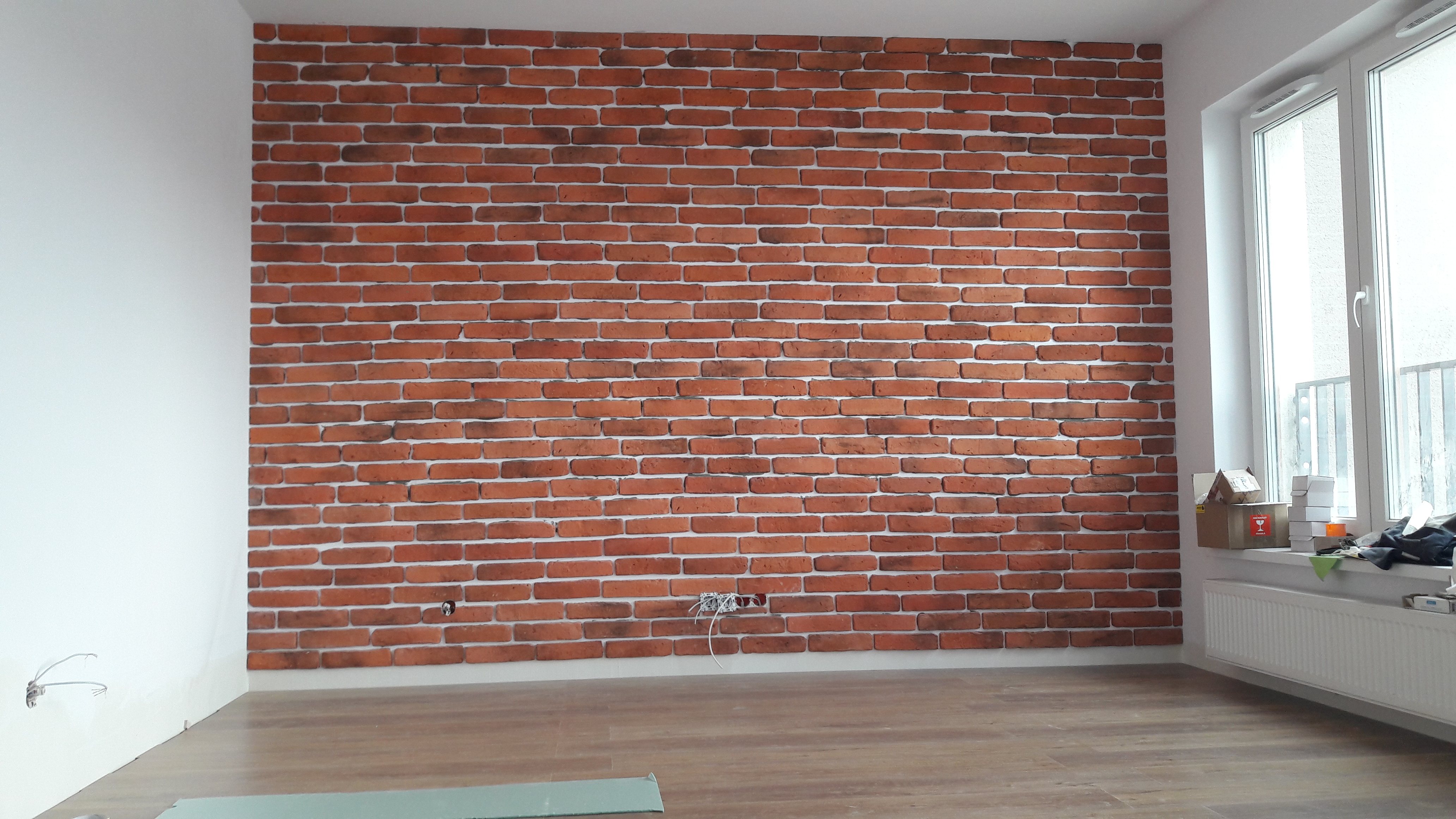 Ściana z cegły dekoracyjnej w nowym mieszkaniu, widoczne kable elektryczne wystające ze ściany, podłoga drewniana, okno z widokiem na balkon.