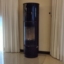 PHU Patryk Szkutnik - Czarny, cylindryczny piec wolnostojący z przeszklonym paleniskiem, umieszczony na przezroczystej płycie ochronnej, na tle jasnych zasłon i beżowej ściany.