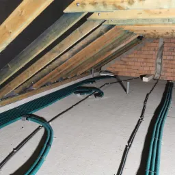 Instalacja przewodów wentylacyjnych w surowym poddaszu z widoczną konstrukcją drewnianą dachu i fragmentem ściany z cegły.