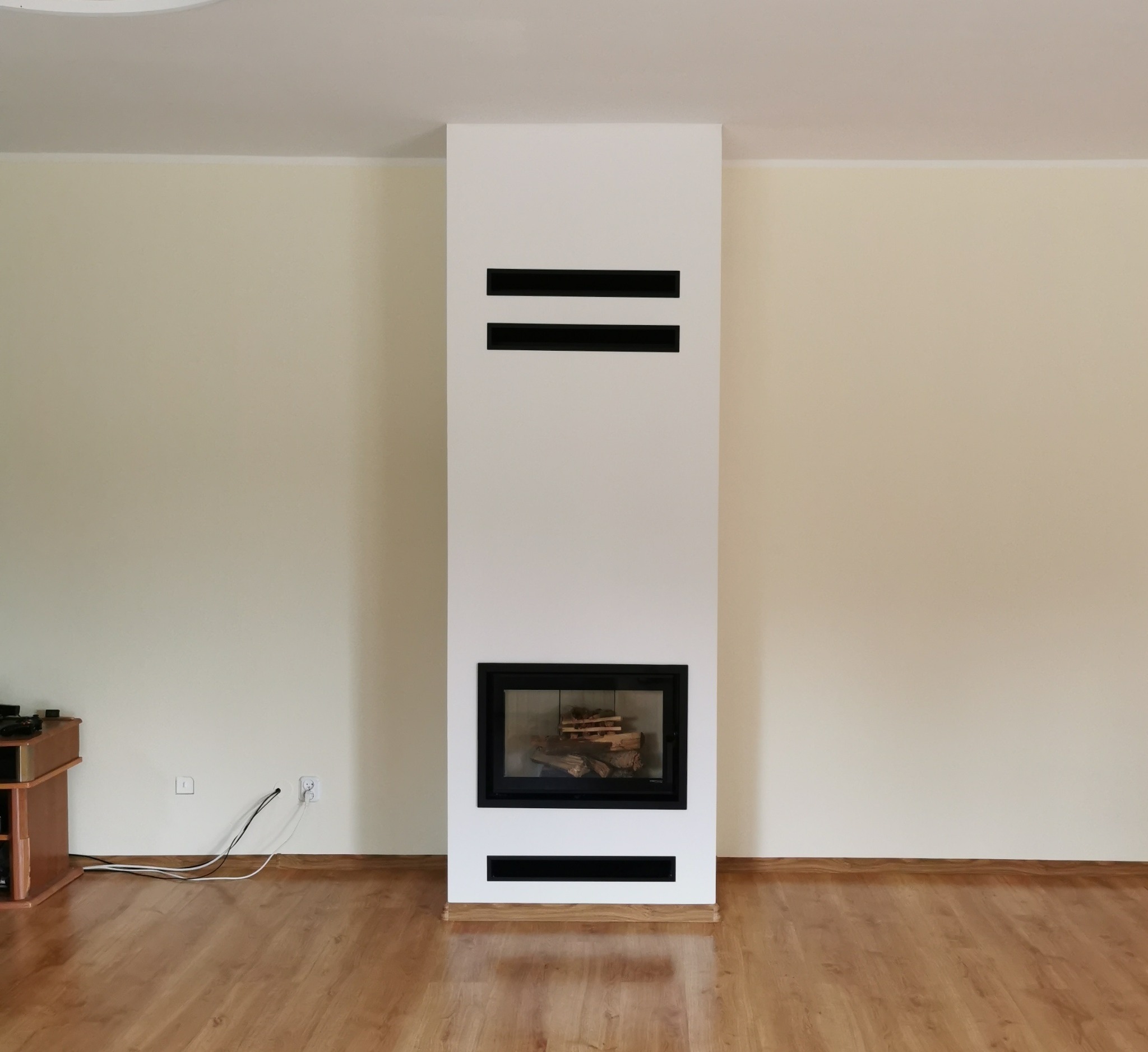 Nowoczesny kominek z czarną ramą, wewnątrz ułożone drewno, dwie poziome czarne szczeliny wentylacyjne w obudowie, jasne ściany i parkiet, widoczne kable od gniazdka.