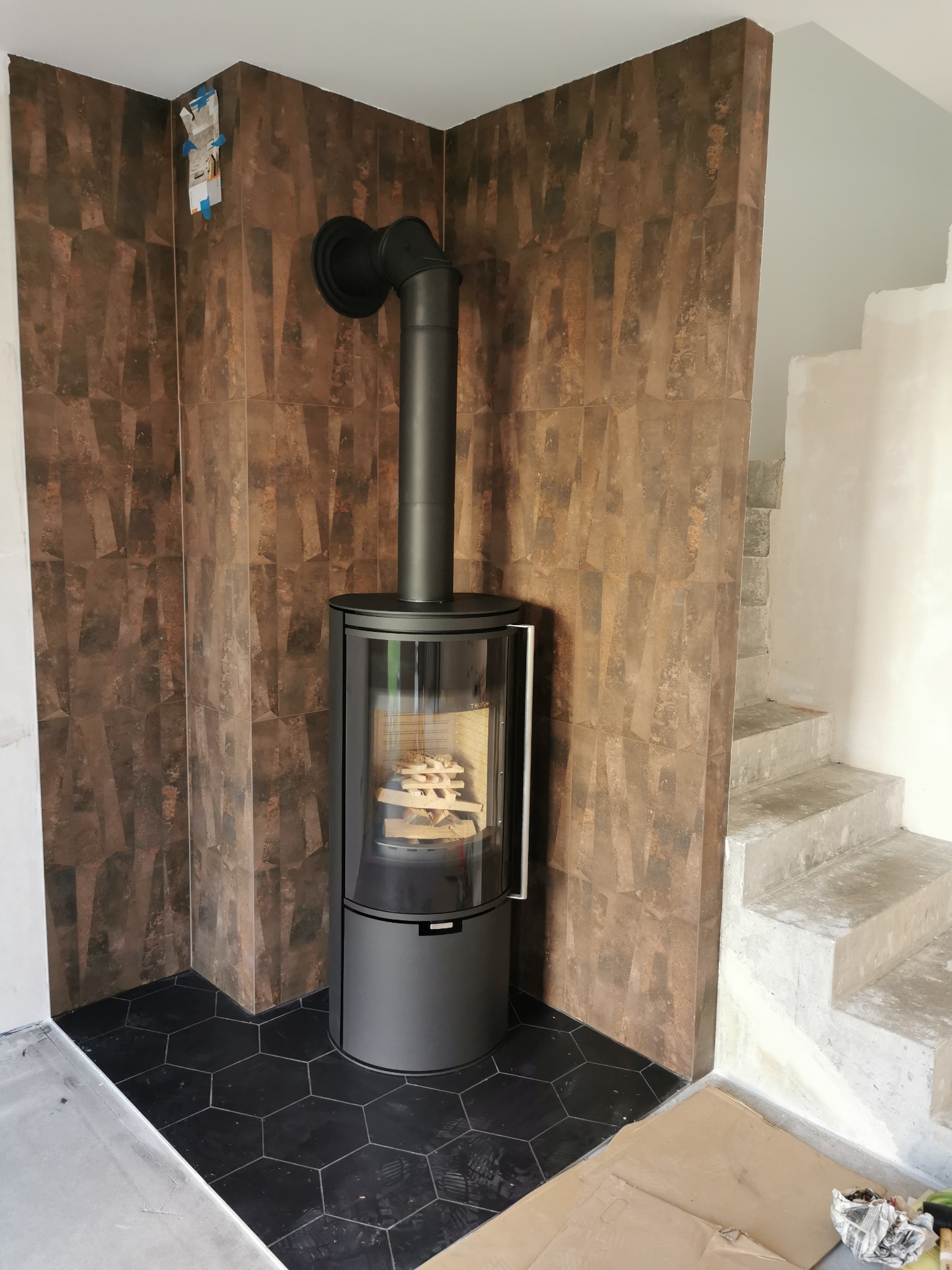 Nowoczesny, czarny piec wolnostojący z przeszklonym frontem, wypełniony drewnem, ustawiony w rogu pomieszczenia wyłożonego brązowymi płytkami imitującymi kamień i czarną heksagonalną podłogą, obok...