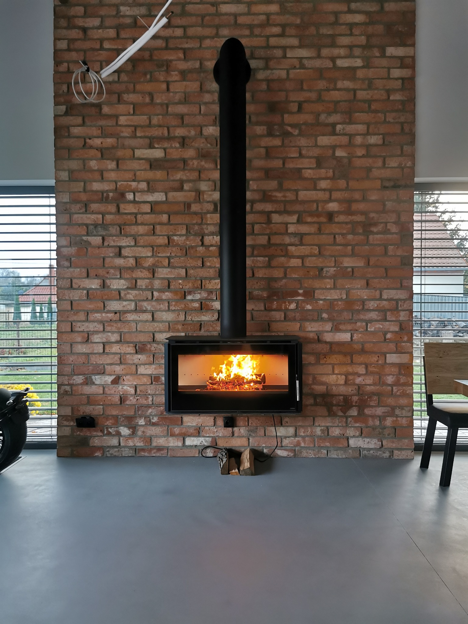 Wnętrze z ceglaną ścianą, na której zawieszony jest czarny, nowoczesny piec z palącym się ogniem. Widoczne drewno obok pieca i fragment motocykla po lewej stronie.