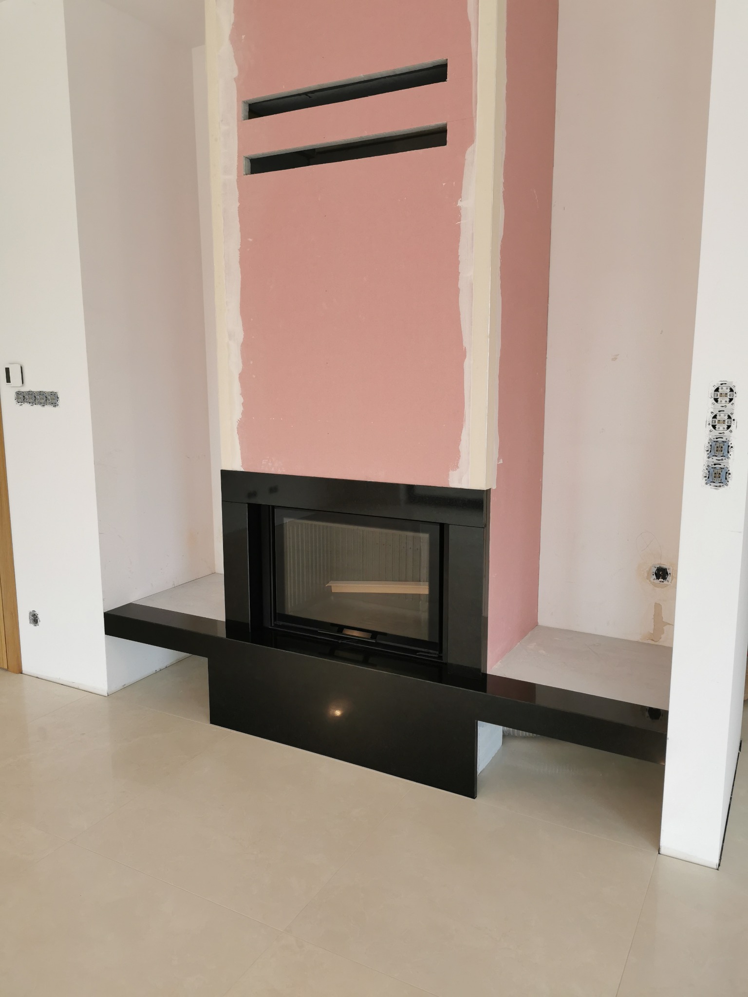 Nowoczesny kominek z czarną obudową w trakcie montażu, widoczne płyty gipsowo-kartonowe i otwory wentylacyjne w obudowie kominka.