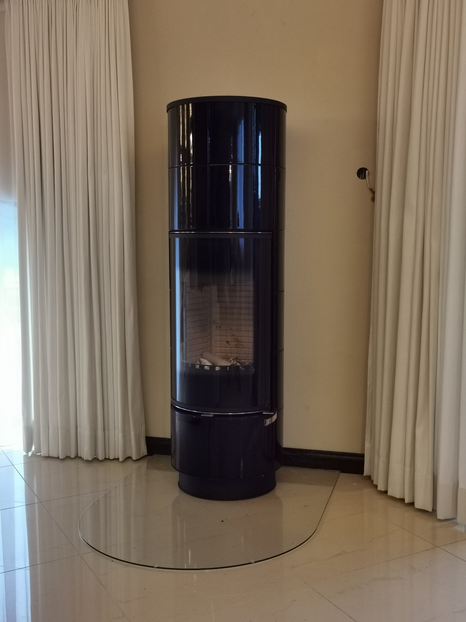 Czarny, cylindryczny piec wolnostojący z przeszklonym paleniskiem, umieszczony na przezroczystej płycie ochronnej, na tle jasnych zasłon i beżowej ściany.