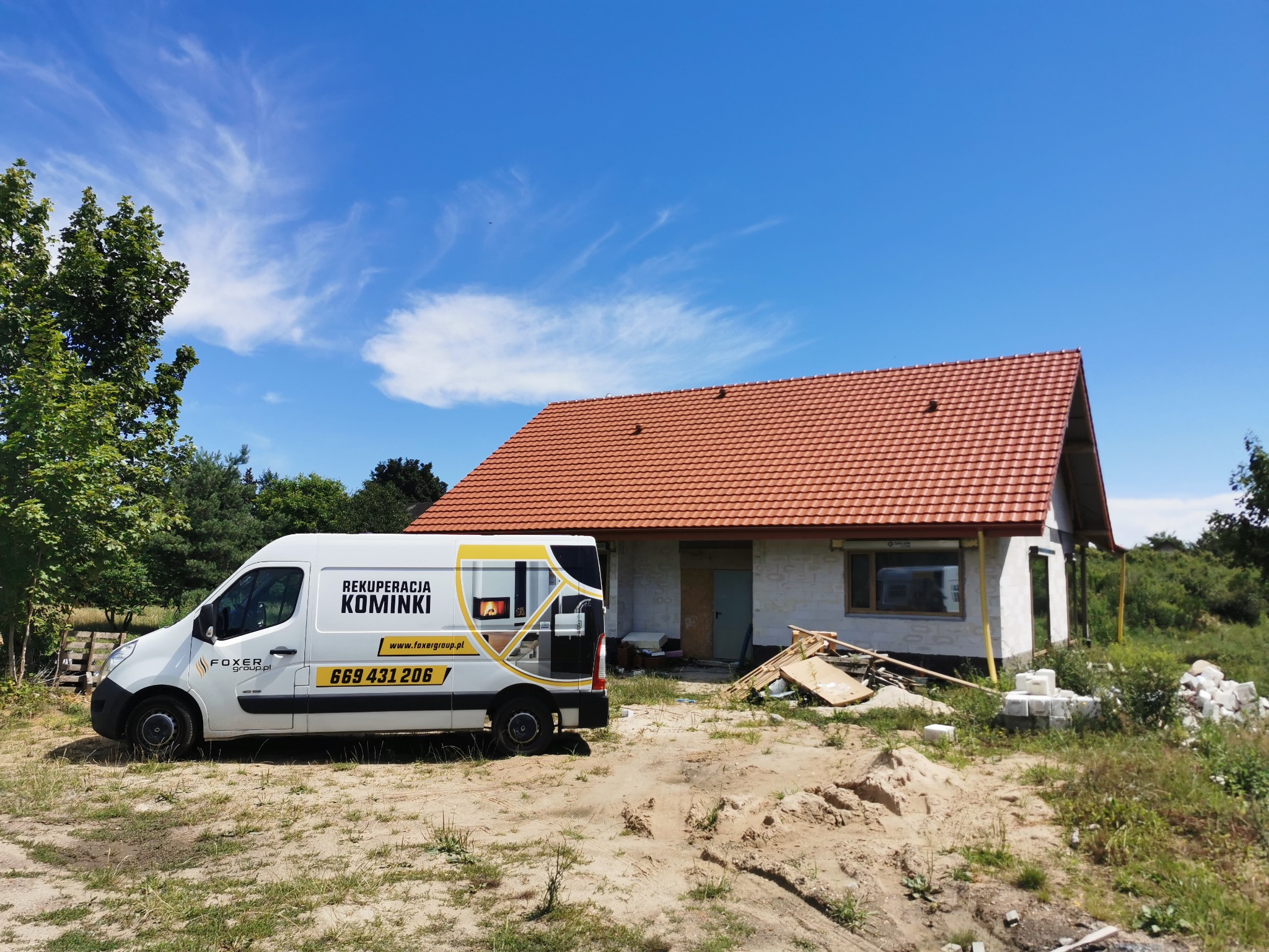 Furgonetka z reklamą firmy instalacyjnej na tle budowanego domu z czerwoną dachówką, widoczne materiały budowlane.