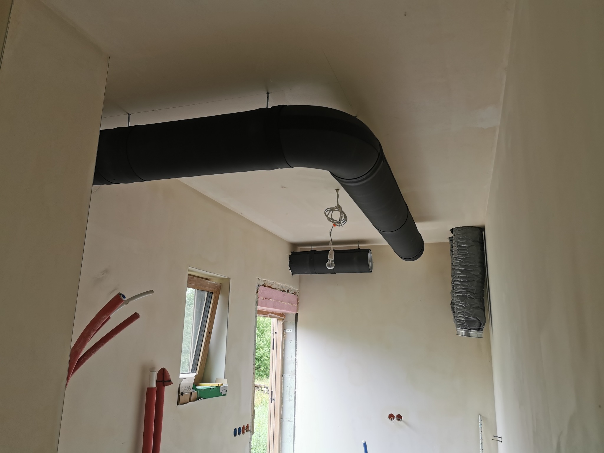 Wnętrze w trakcie prac wykończeniowych z widocznymi elementami instalacji wentylacyjnej: czarnymi izolowanymi rurami pod sufitem, elastycznym przewodem wentylacyjnym oraz wystającymi rurami...