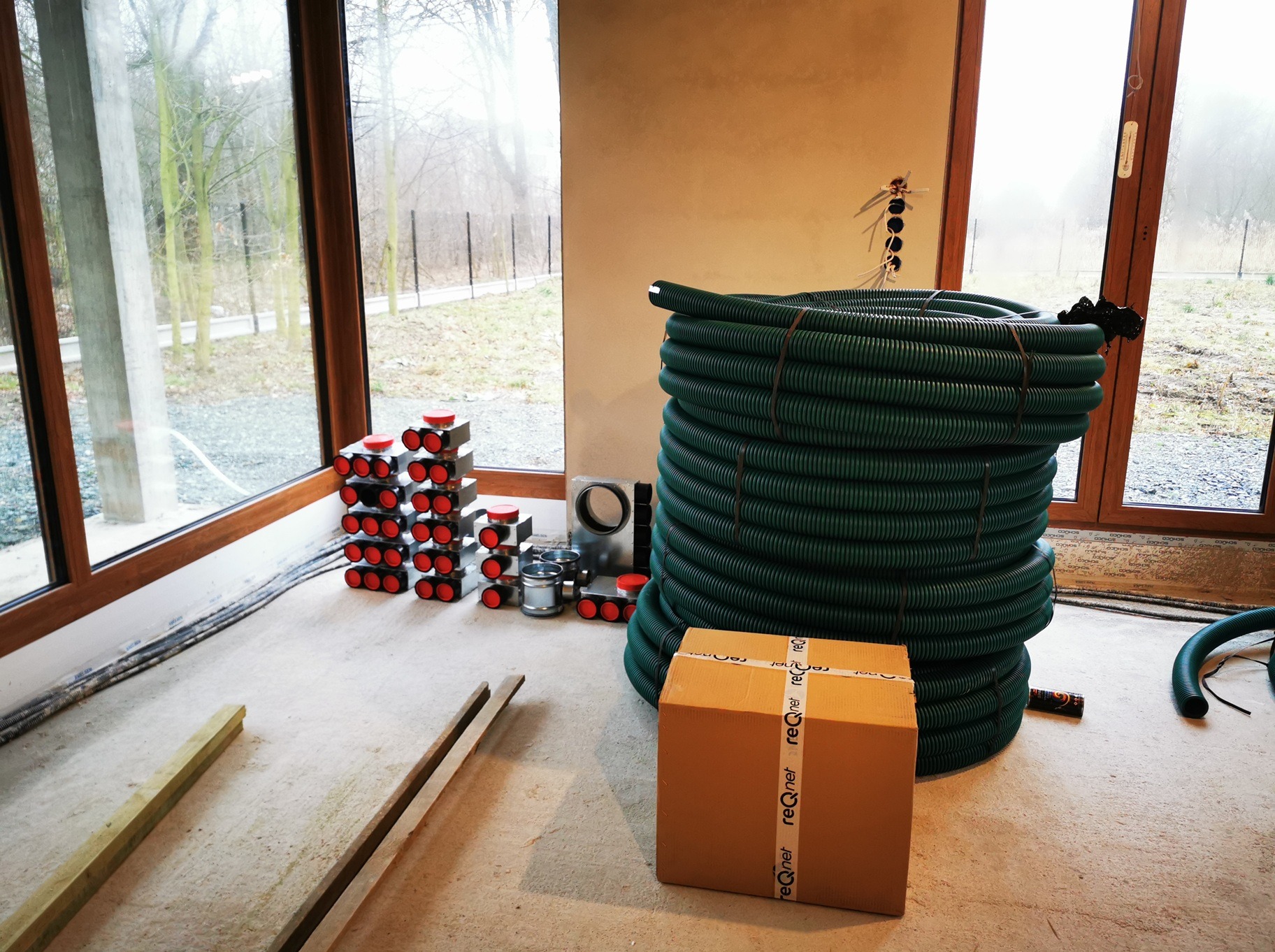 Stos zielonych rur rekuperacyjnych, skrzynka z napisem 'reqnet' i rozdzielacze powietrza z czerwonymi pokrywami w surowym wnętrzu z dużymi oknami