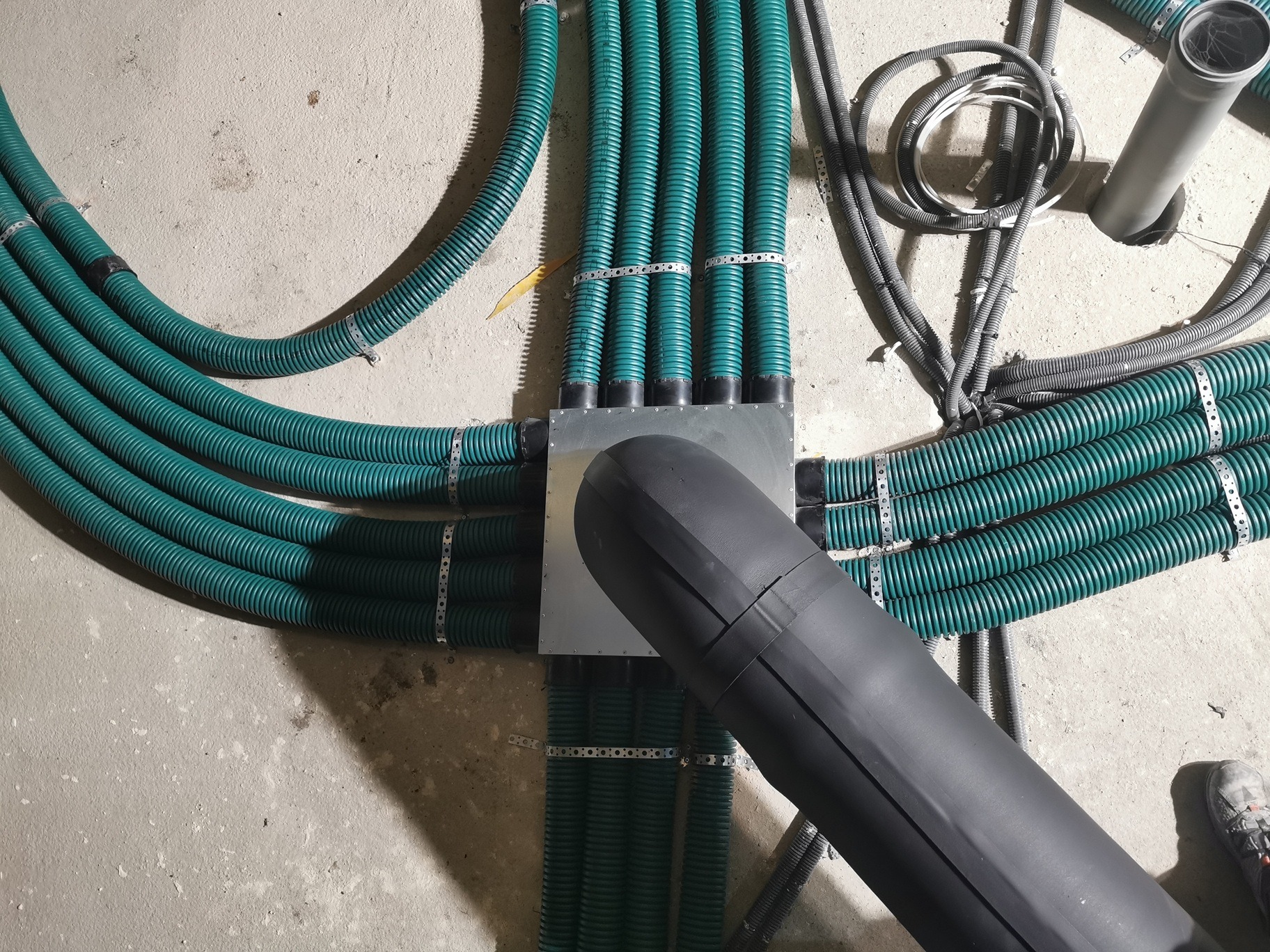 Rozprowadzona instalacja wentylacyjna z zielonych, karbowanych rur, połączonych metalową puszką i czarnym przewodem, widoczne metalowe obejmy mocujące, surowy beton na podłodze, but.