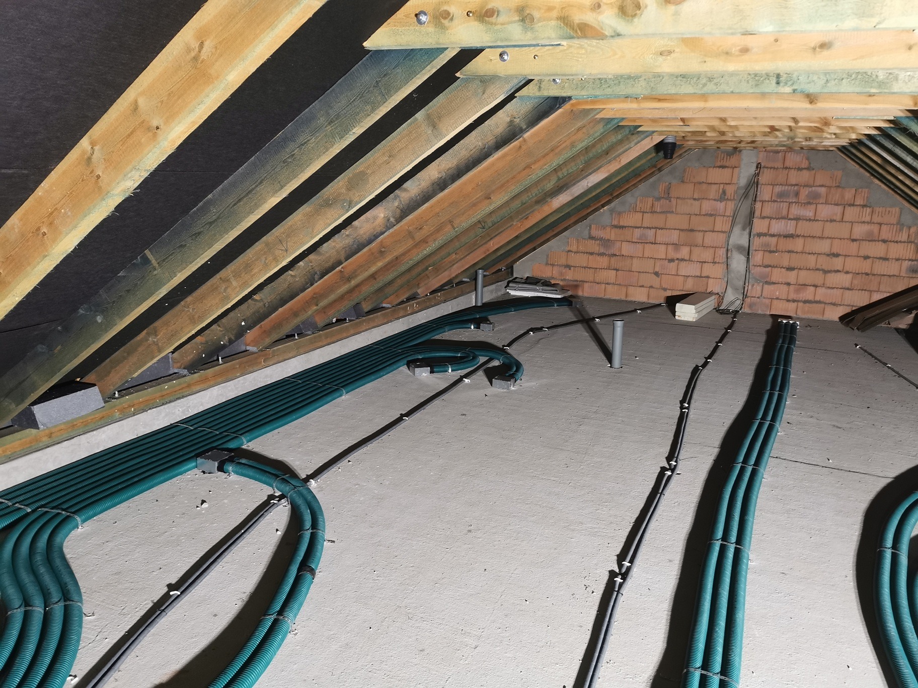 Instalacja przewodów wentylacyjnych w surowym poddaszu z widoczną konstrukcją drewnianą dachu i fragmentem ściany z cegły.