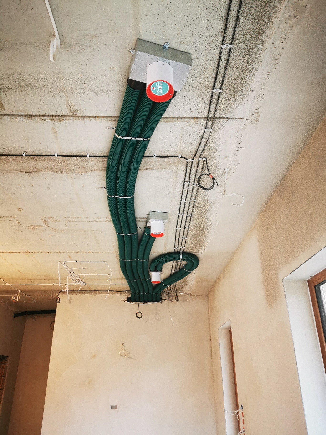 Zielone, elastyczne przewody wentylacyjne zamontowane pod betonowym stropem w surowym wnętrzu, widoczne także kable elektryczne i fragment okna.