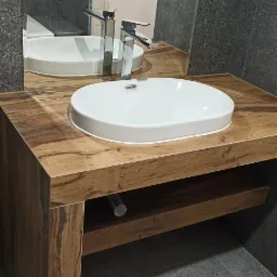 Umywalka nablatowa owalna, biała, z chromowaną baterią, na blacie z drewna z półką, szare płytki na ścianie, fragment instalacji elektrycznej widoczny.