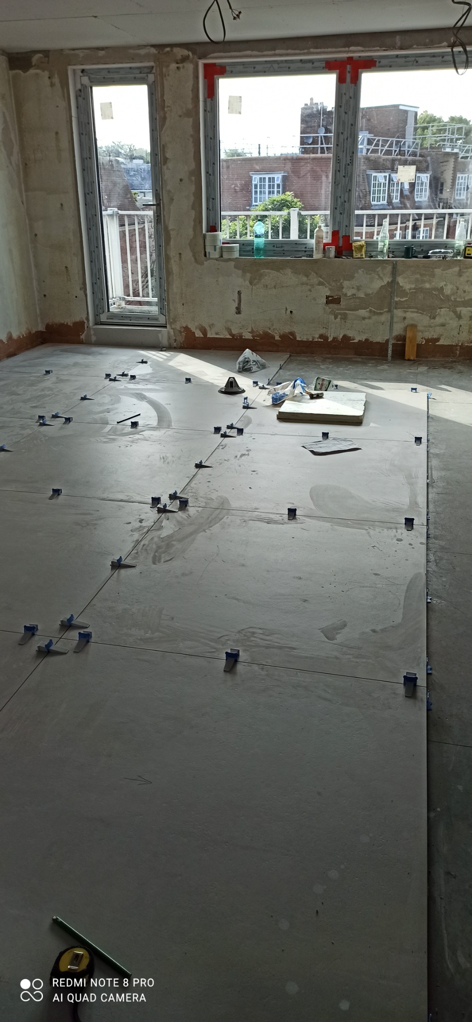 Układanie szarych płytek podłogowych z użyciem niebieskich klipsów poziomujących, widok na okna i materiały budowlane w tle.