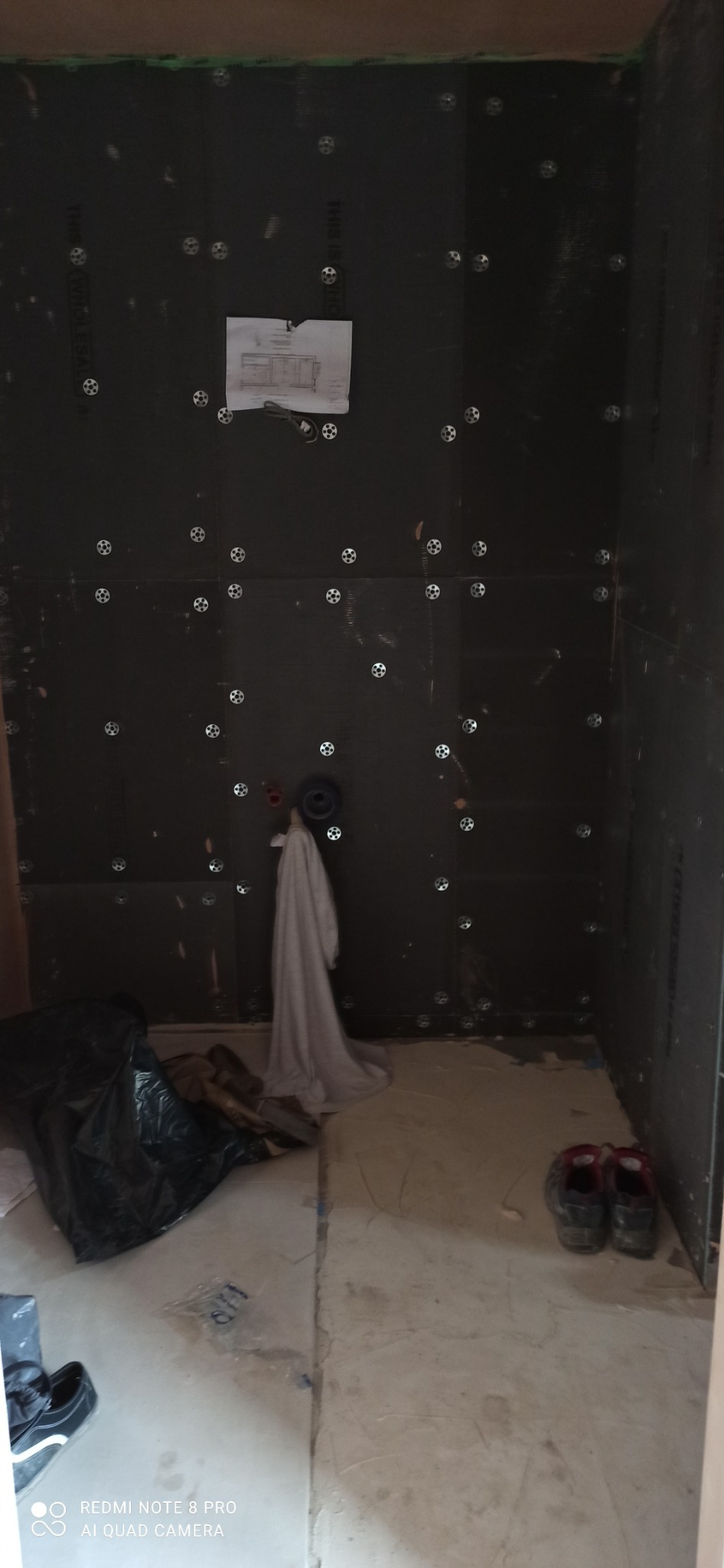 Przygotowanie do układania płytek w małej łazience: czarna płyta izolacyjna z kołkami mocującymi na ścianach, wystająca rura instalacji wodnej, projekt łazienki przyklejony do ściany, podłoga...