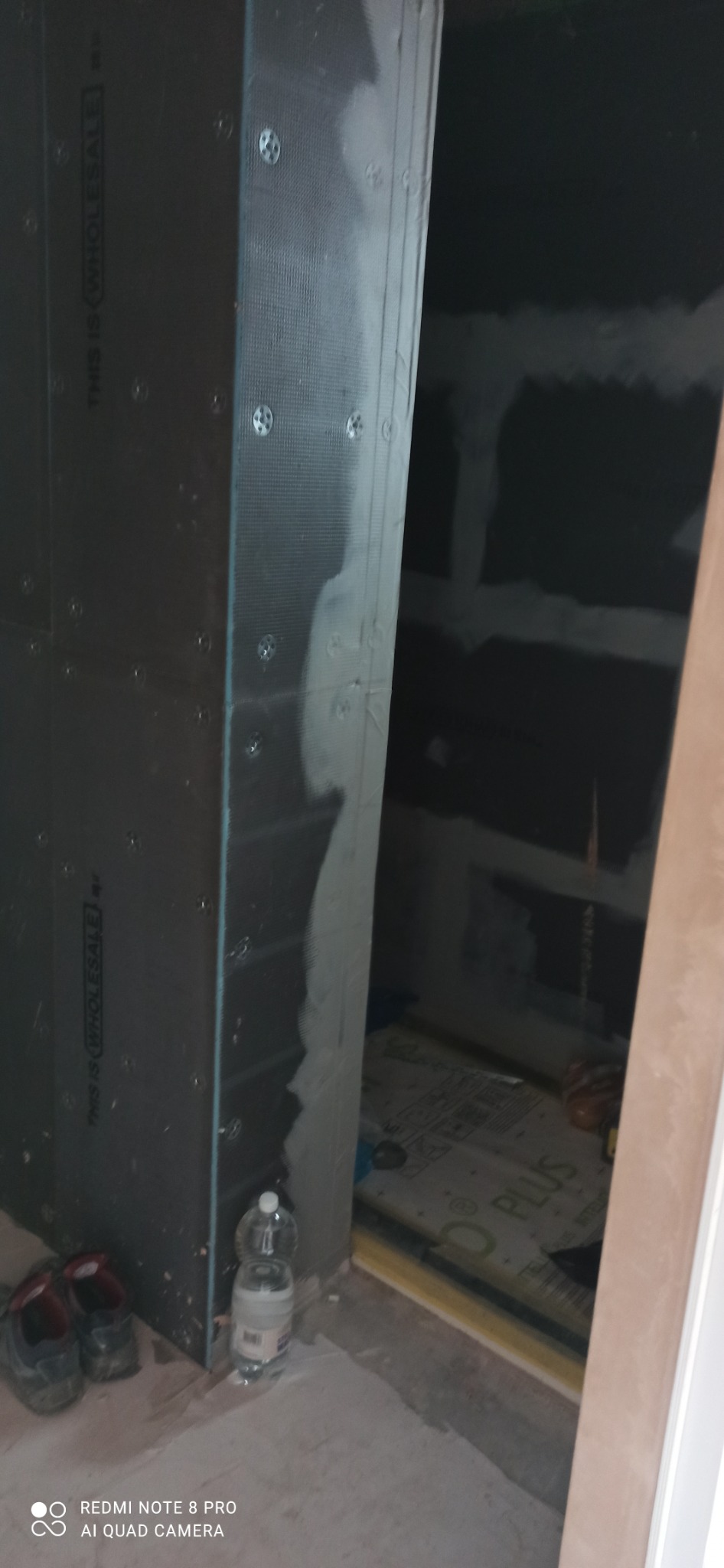 Montaż płyty karton gips w narożniku pomieszczenia, widoczne mocowania, częściowo zaszpachlowane łączenia, w tle przygotowane ściany do dalszych prac, na podłodze leżą buty robocze i butelki z wodą.