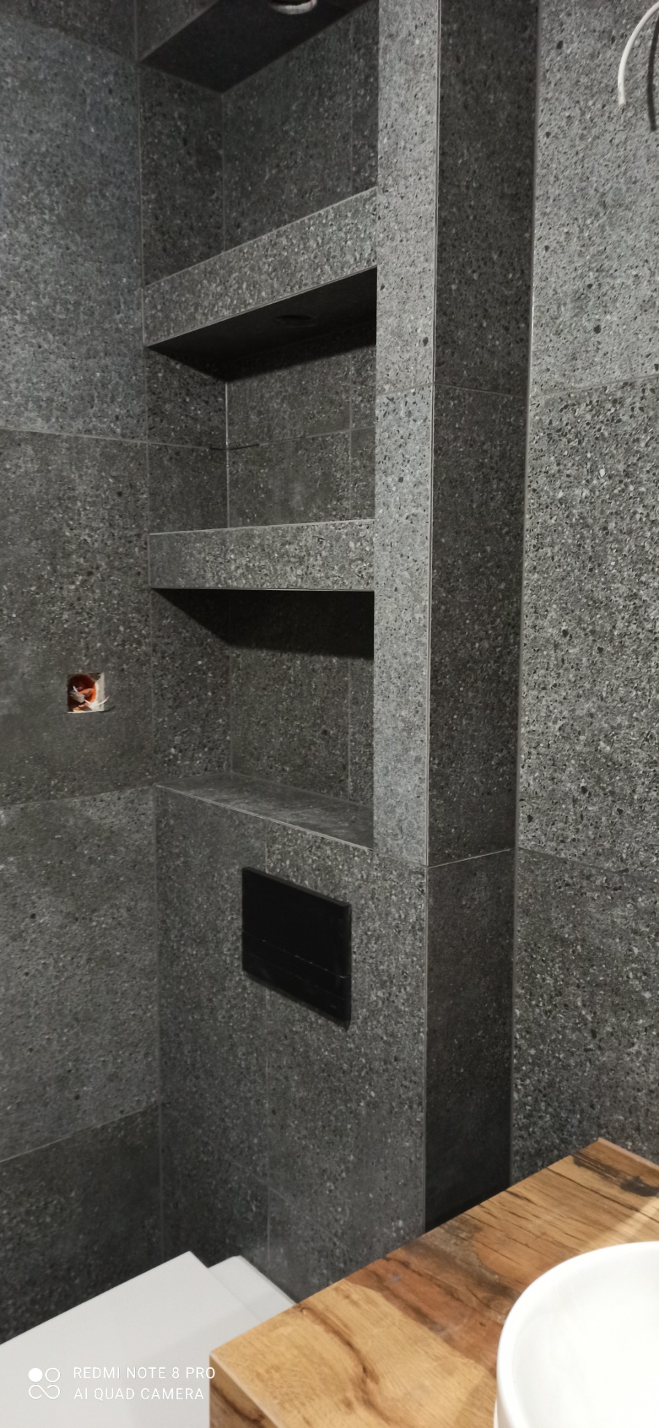 Nowoczesna łazienka z szarymi płytkami imitującymi kamień, wnęki w ścianie wykończone tym samym materiałem, czarny panel sterujący spłuczką, fragment drewnianego blatu z umywalką.
