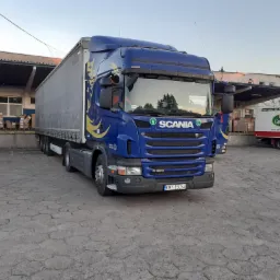 Niebieska ciężarówka Scania G420 z naczepą, z dekoracyjnymi złotymi elementami, zaparkowana na placu w Skomielnej Białej, w tle budynek magazynowy z rampą i częściowo widoczna ciężarówka z logo...