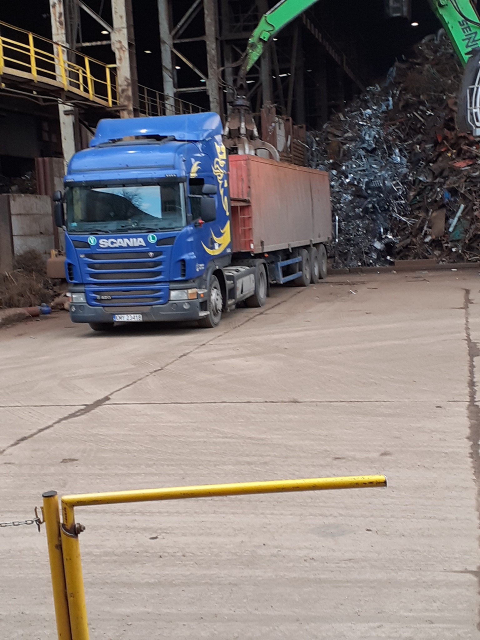 Niebieska ciężarówka Scania z pomarańczową naczepą na betonowym placu, w tle sterta złomu ładowana przez zielony dźwig.