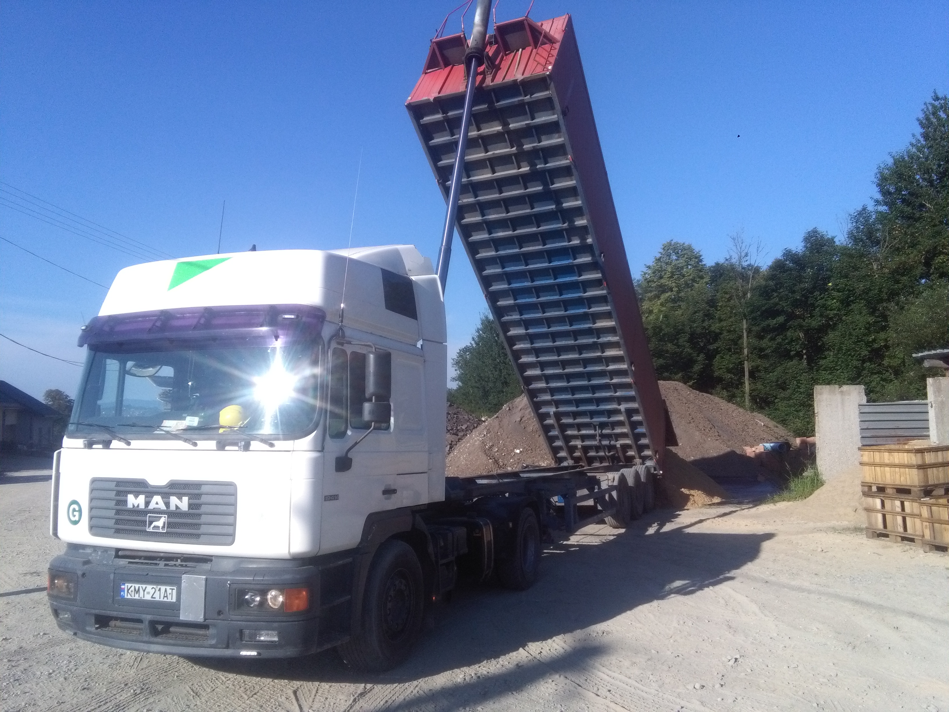 Ciężarówka MAN z podniesioną czerwoną skrzynią ładunkową, wysypująca piasek na placu budowy w słoneczny dzień.