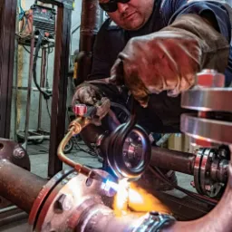 Pracownik w okularach ochronnych i rękawicach spawalniczych łączy rury z kołnierzami za pomocą palnika acetylenowo-tlenowego, widoczne iskry i płomień, w tle konstrukcja metalowa i butla z gazem.