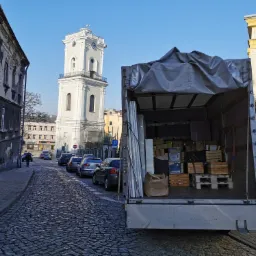 Otwarta paka samochodu dostawczego załadowana kartonami i drewnianymi skrzynkami na paletach, w tle brukowana ulica i wieża zegarowa.