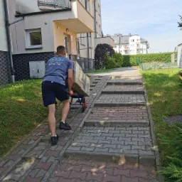 Pracownik firmy transportowej w szarej koszulce z logo na plecach, wciąga wózek z zapakowanym kartonem po rampie dla wózków inwalidzkich obok schodów prowadzących do bloku mieszkalnego.