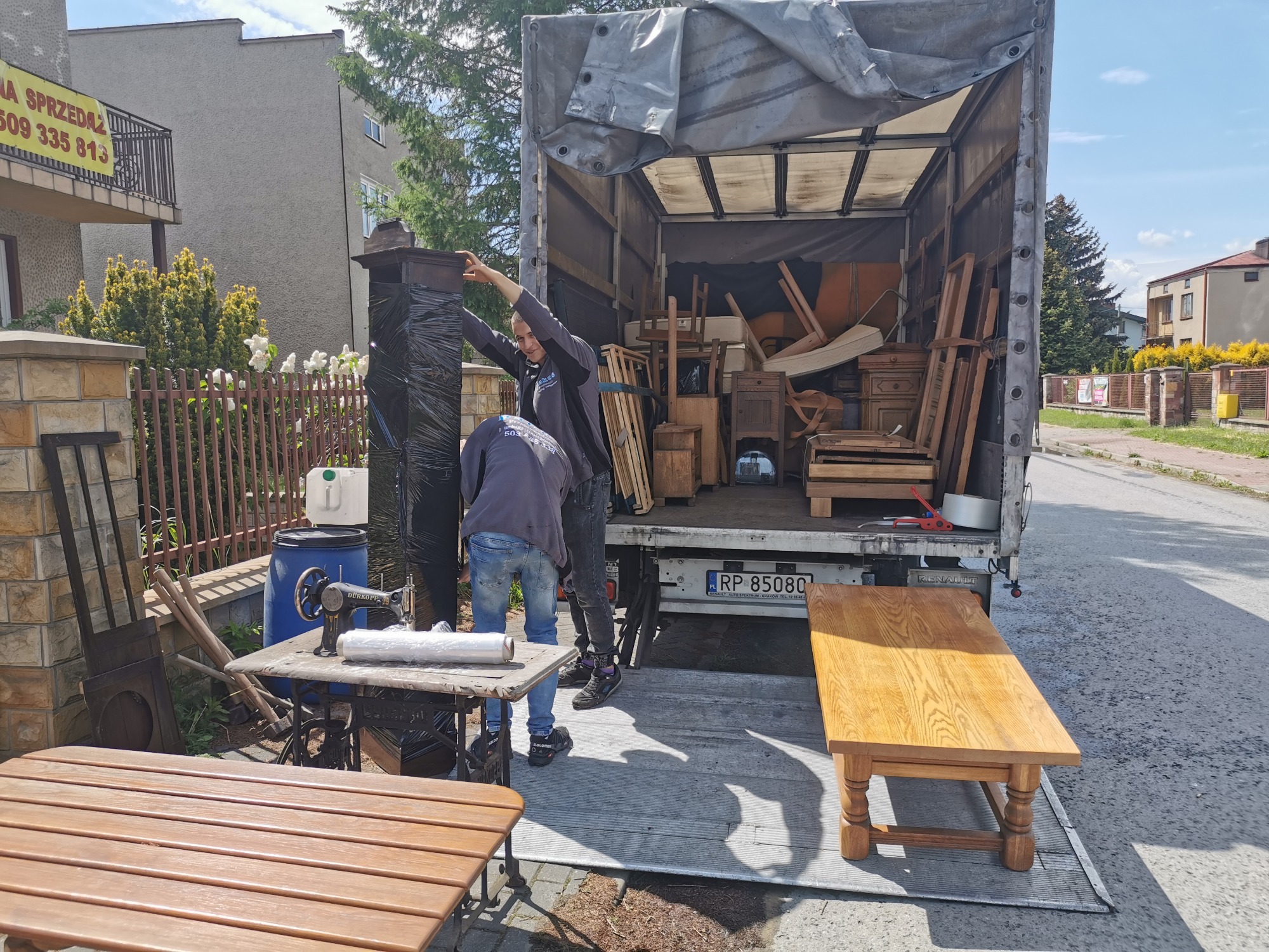 Załadunek mebli, w tym krzeseł, stołów i szafek, do ciężarówki podczas przeprowadzki, widoczny również stary stół z maszyną do szycia.