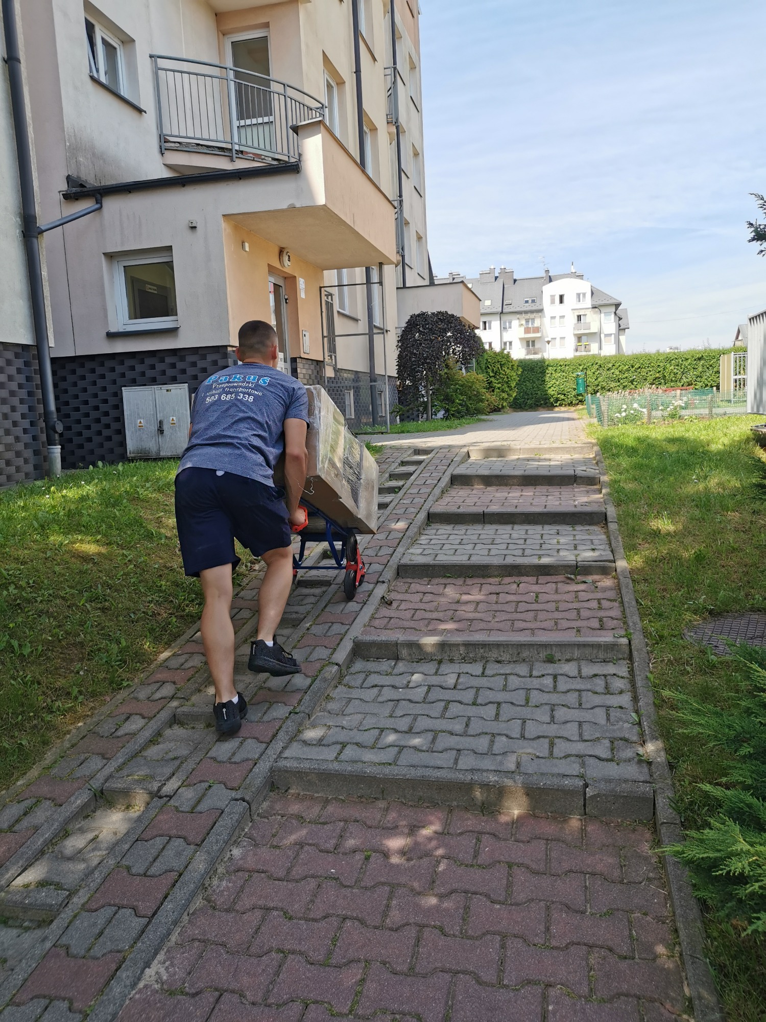 Pracownik firmy transportowej w szarej koszulce z logo na plecach, wciąga wózek z zapakowanym kartonem po rampie dla wózków inwalidzkich obok schodów prowadzących do bloku mieszkalnego.